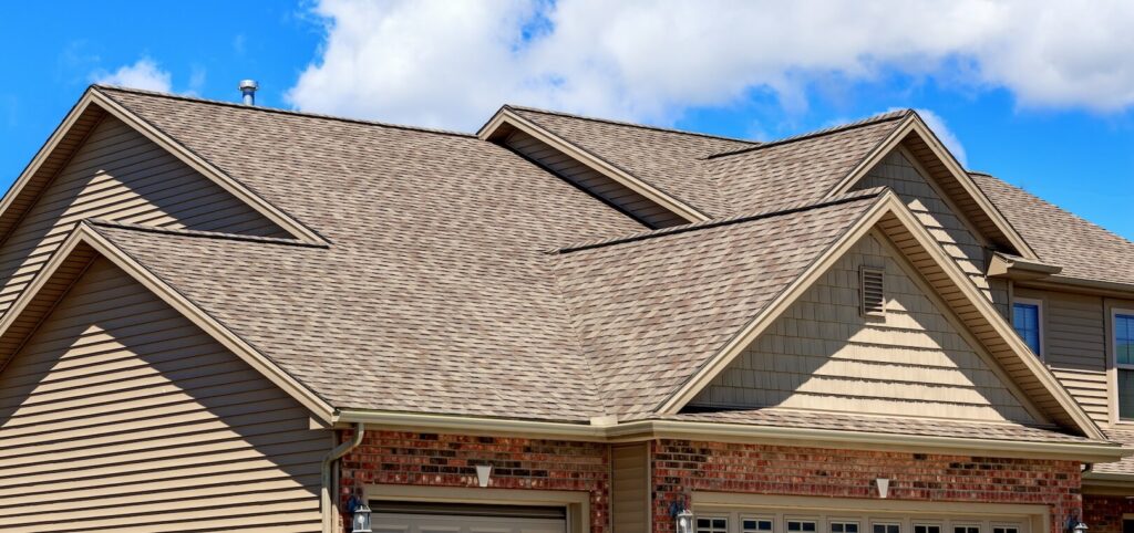 Roof and upper facade of a suburban house with beige vinyl siding and multiple asphalt shingle-covered gables under a clear blue sky.