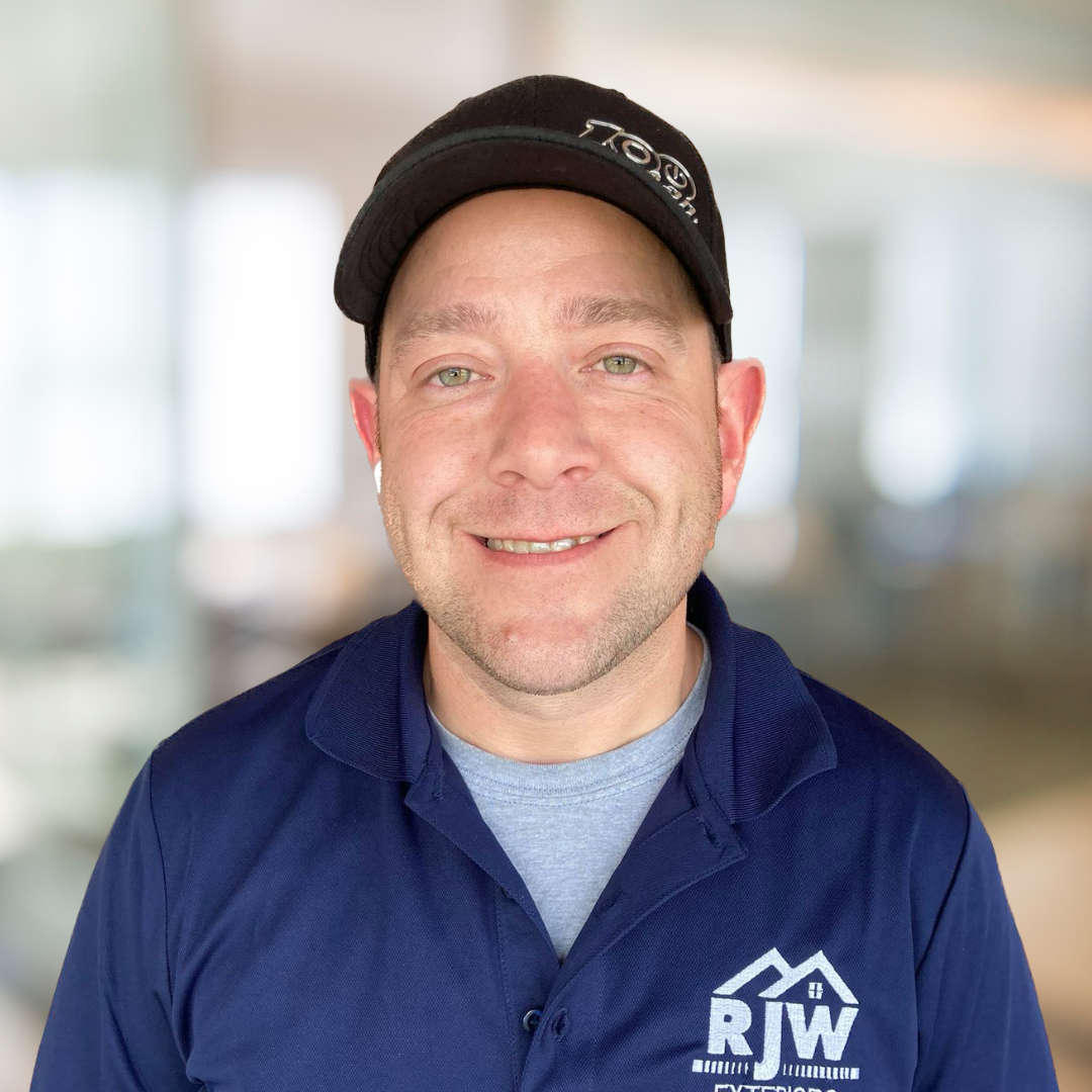 A man wearing a black cap and a navy blue polo shirt with an "RW" logo is smiling at the camera. The background is a blurred indoor setting.