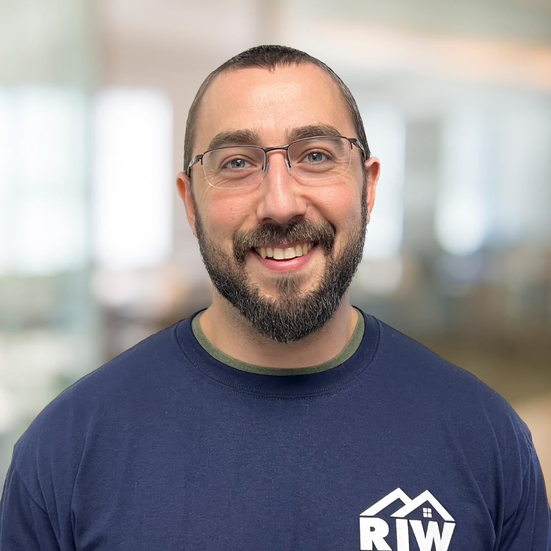 A man with a beard and glasses smiles at the camera. He is wearing a blue shirt with the letters "RIW" on it. The background is blurred and appears to be an indoor setting.