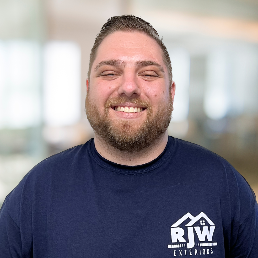 A person wearing a navy blue RJW Exteriors t-shirt is smiling with a blurred indoor background.