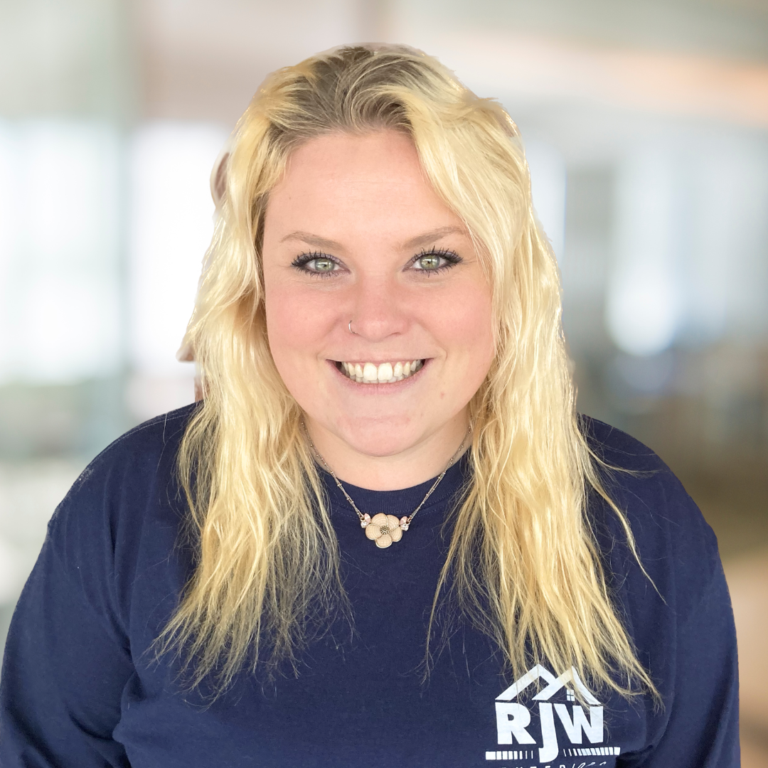 A person with long blonde hair smiles at the camera, wearing a dark shirt with a logo that reads "RJN". The background is blurred.