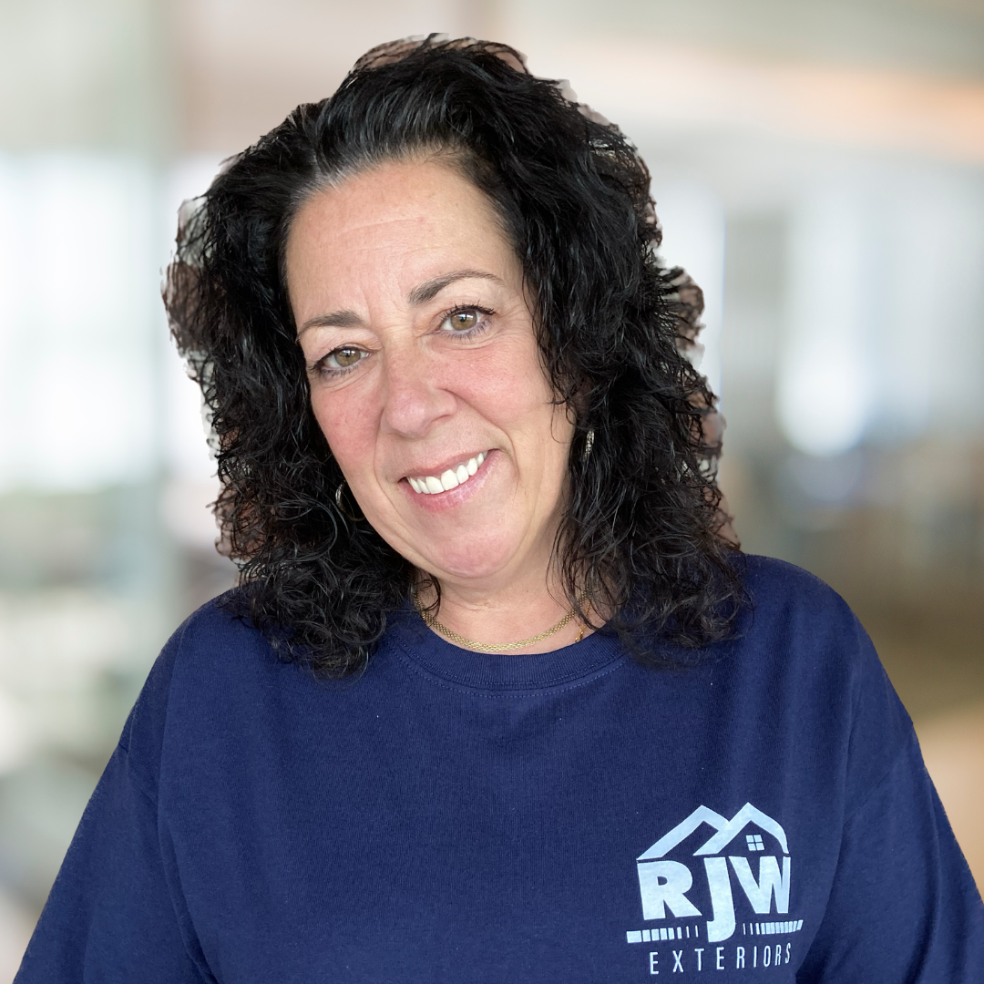 A person with curly dark hair wearing a navy blue shirt with an "RJW Exteriors" logo smiles at the camera. The background appears to be an indoor space with a blurred setting.
