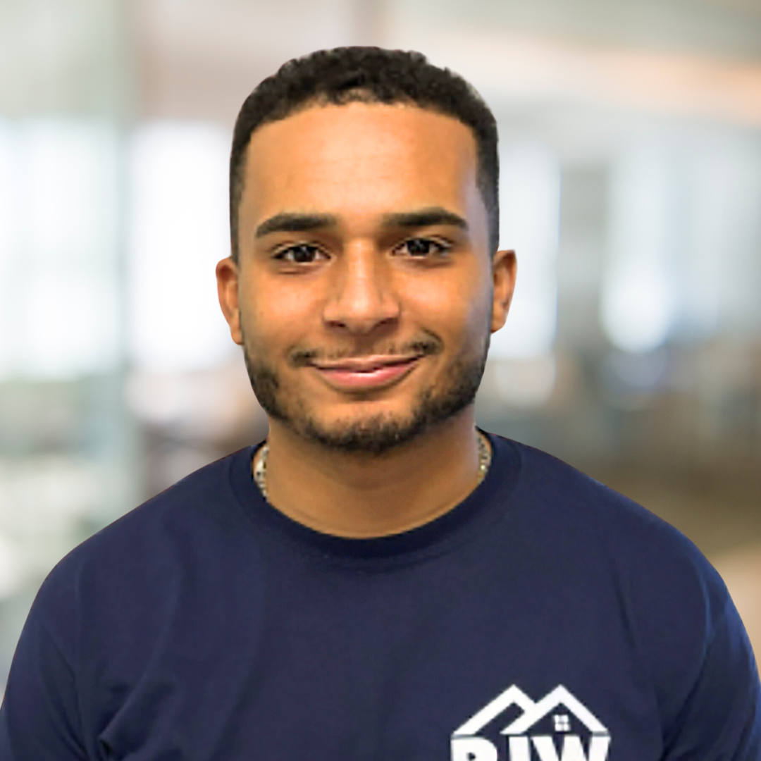 Young man with short curly hair and a trimmed beard, wearing a navy blue shirt with a white logo. The background is blurred, suggesting an indoor setting, possibly an office or workspace.