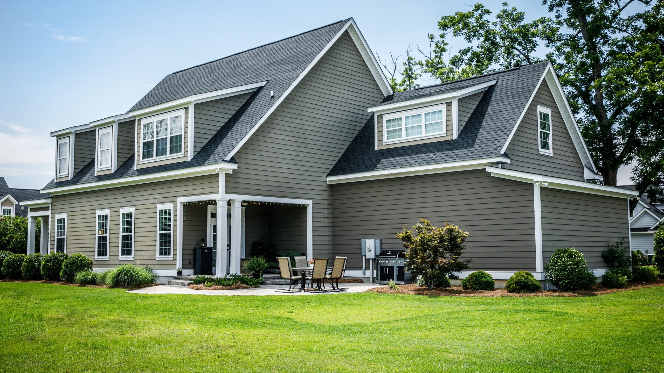 Two-story gray house with dormers, covered patio, and lush lawn on a sunny day.