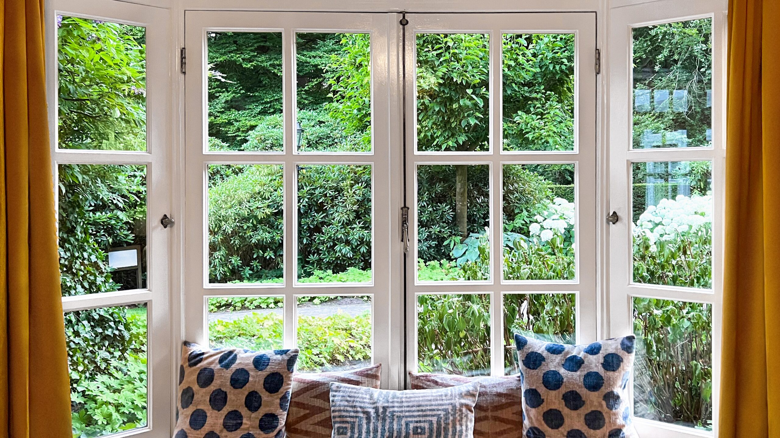 Interior view of a large bay window framed by curtains overlooking a lush outdoor scene