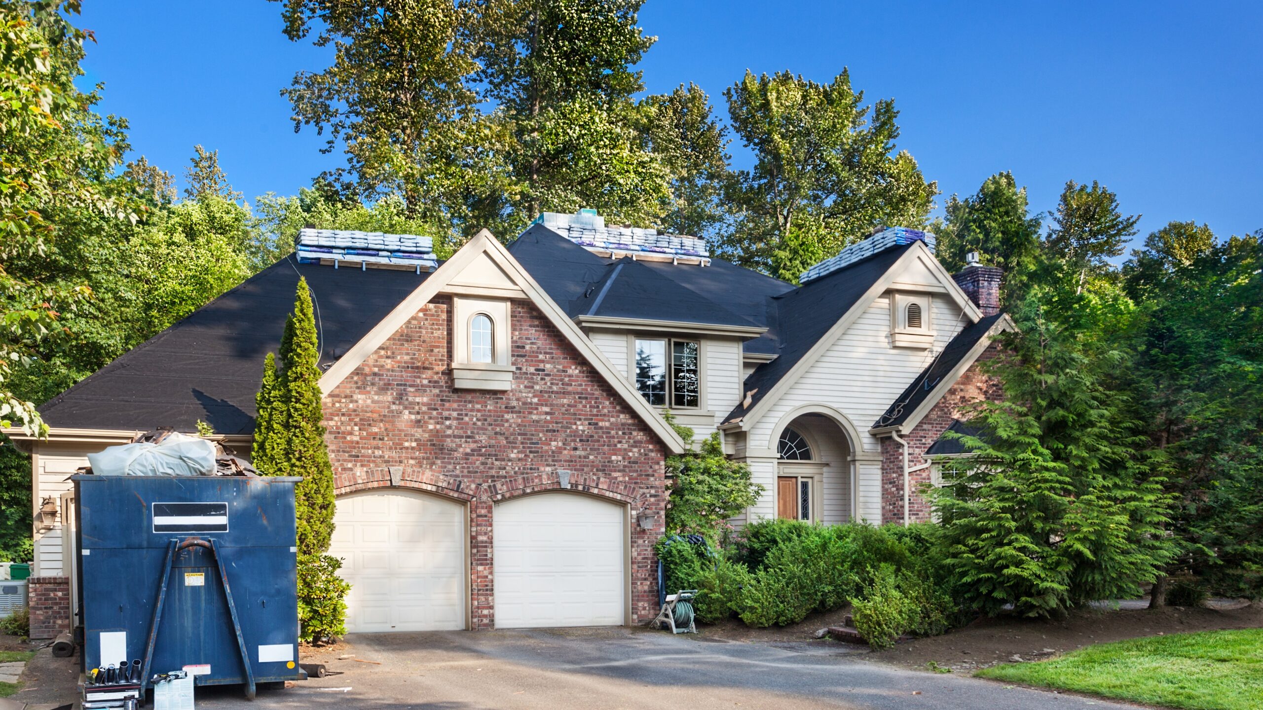 A large suburban home with old shingles stowed in a dumpster and stacks of new shingles on its roof in preparation for a full replacement